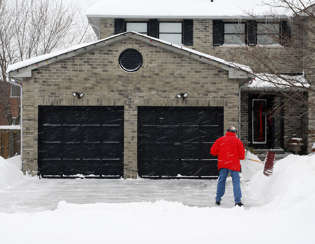Man in a red jacket shoveling snow from a driveway in front of a two car brick garage