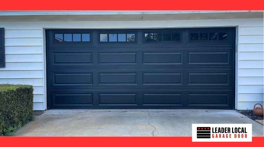 Dark gray sectional garage door with small rectangular windows along the top row, set in white siding with a concrete driveway and a hedge on the left.
